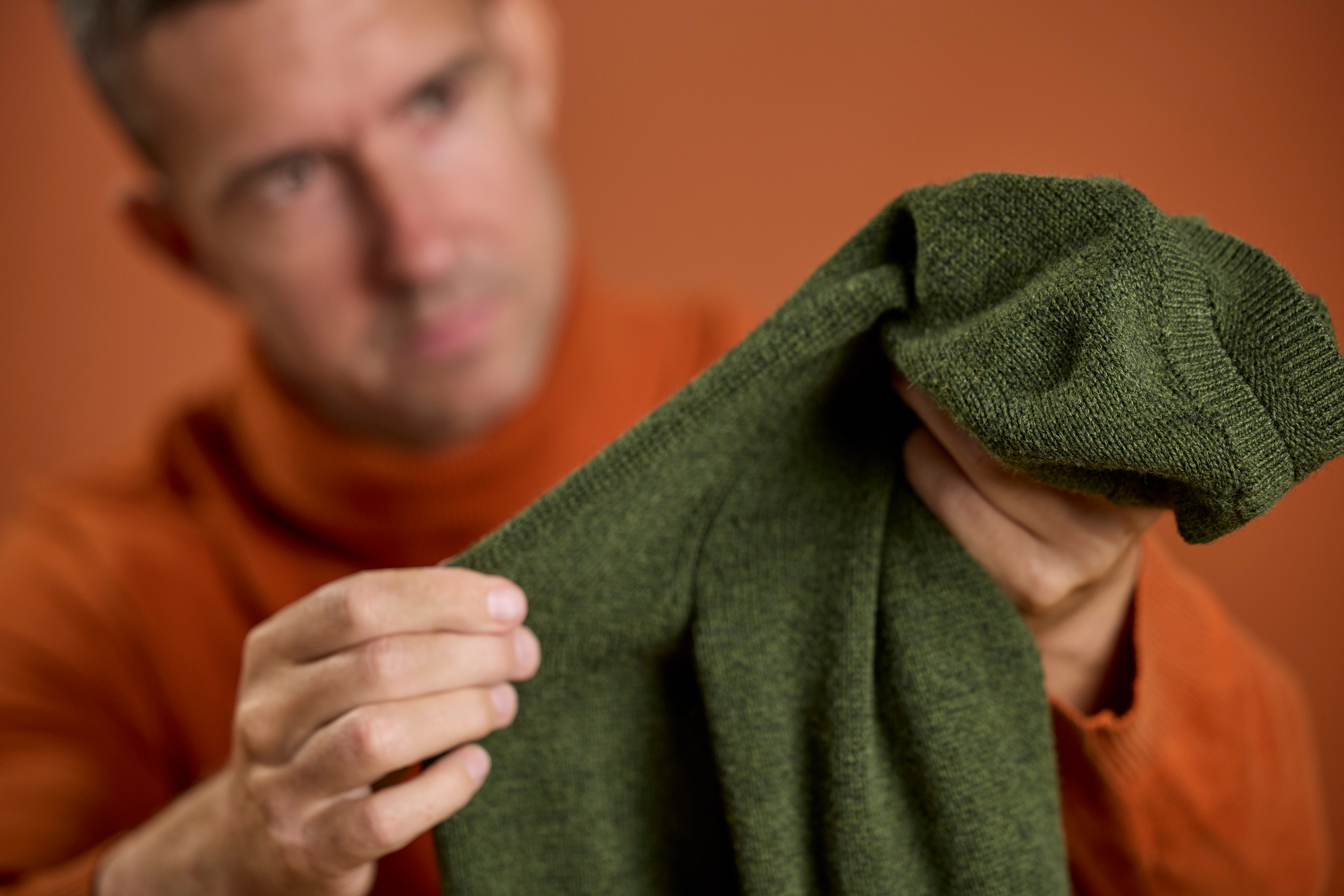 En mann som skeptisk inspiserer et grønt klesplagg, stående mot en oransje bakgrunn.