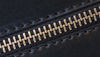 A close-up image of a black faux leather texture featuring a metallic zipper with gold teeth.