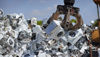 A claw crane lifts two scrap washing machines from a large pile of electronic waste in a scrap yard.