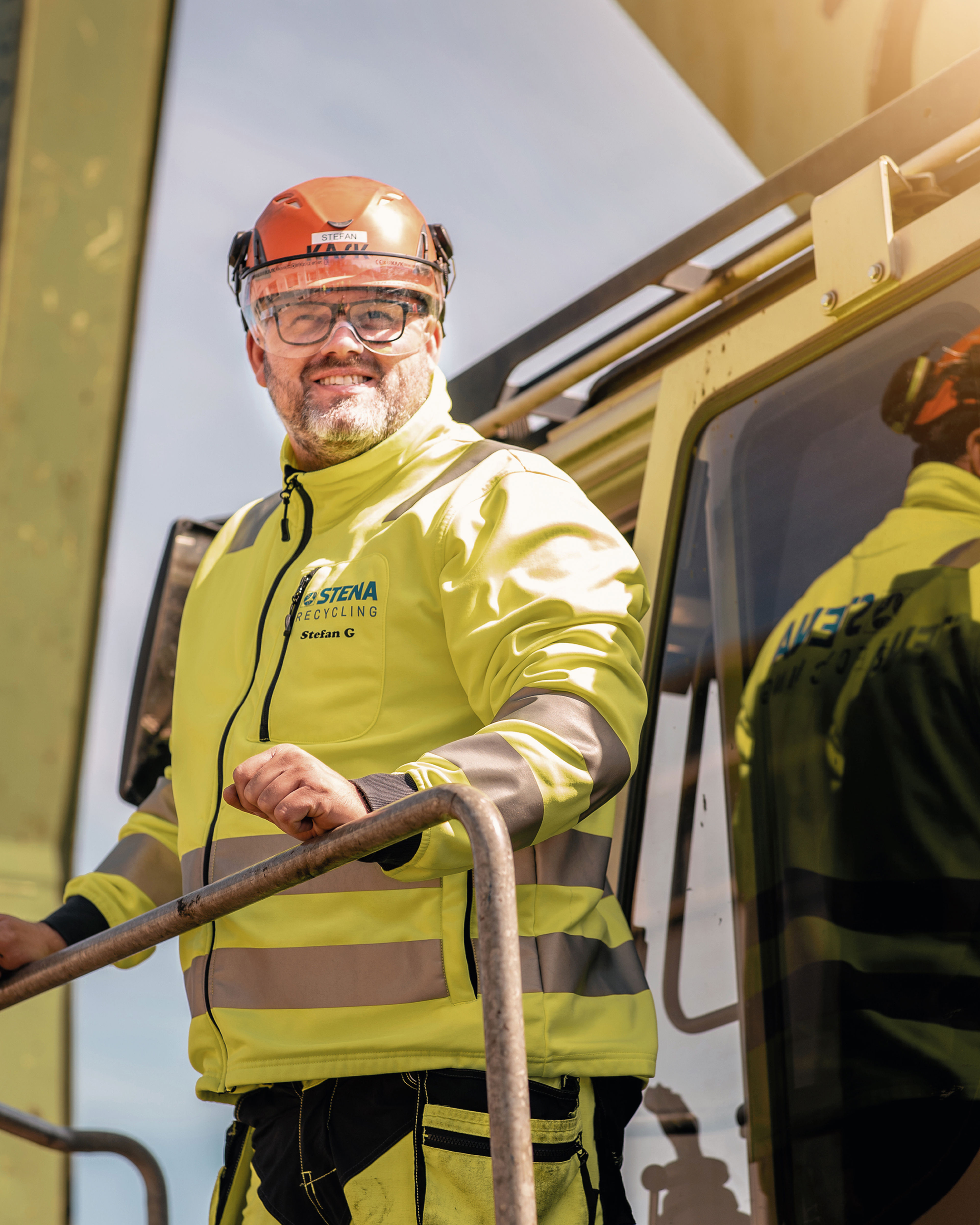 Male Stena Recycling expert wearing a hardhat and reflective work jacket, climbing out of a yellow vehicle