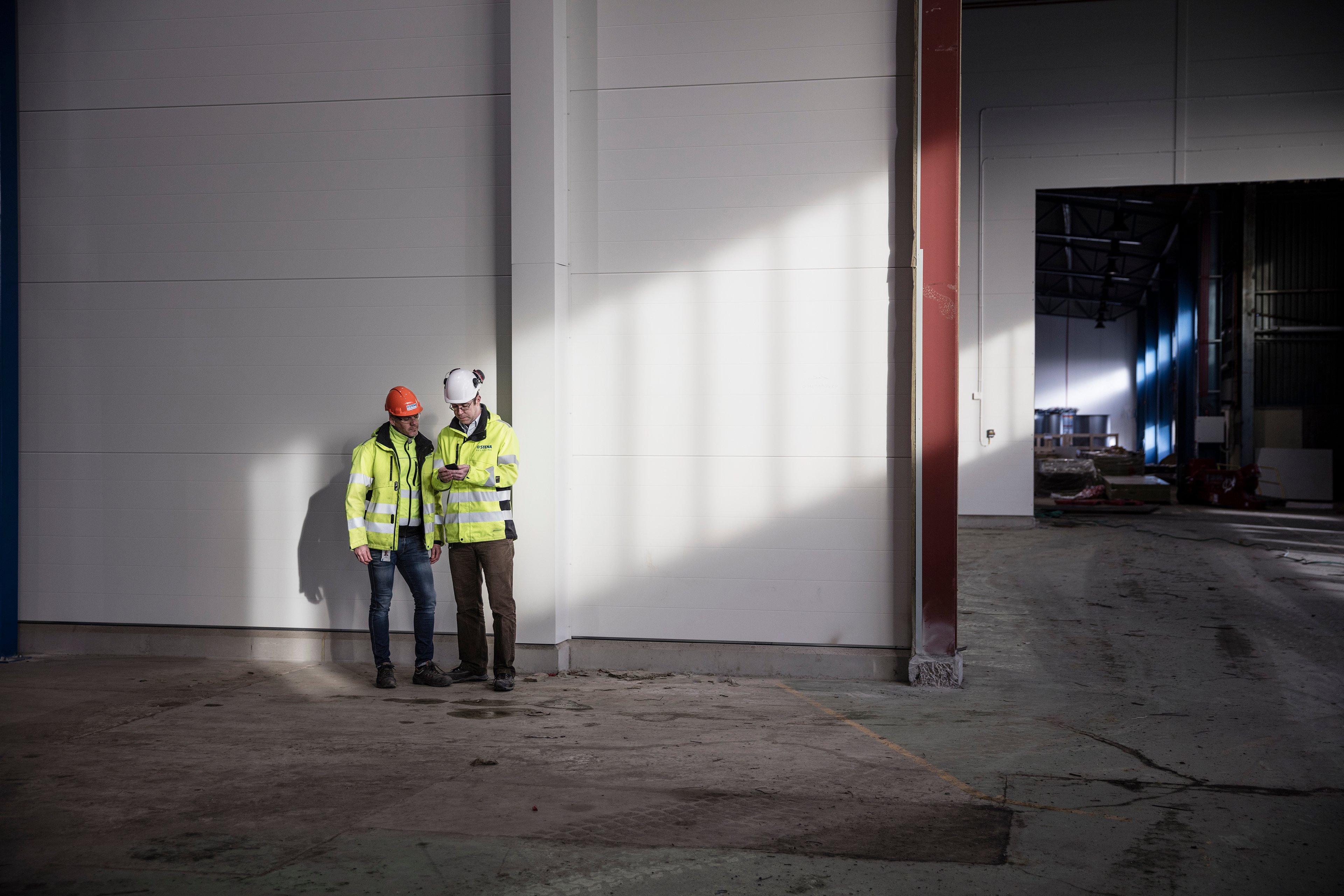 Two male Stena Recycling employees in protective gear look at a mobile phone together in a recycling facility