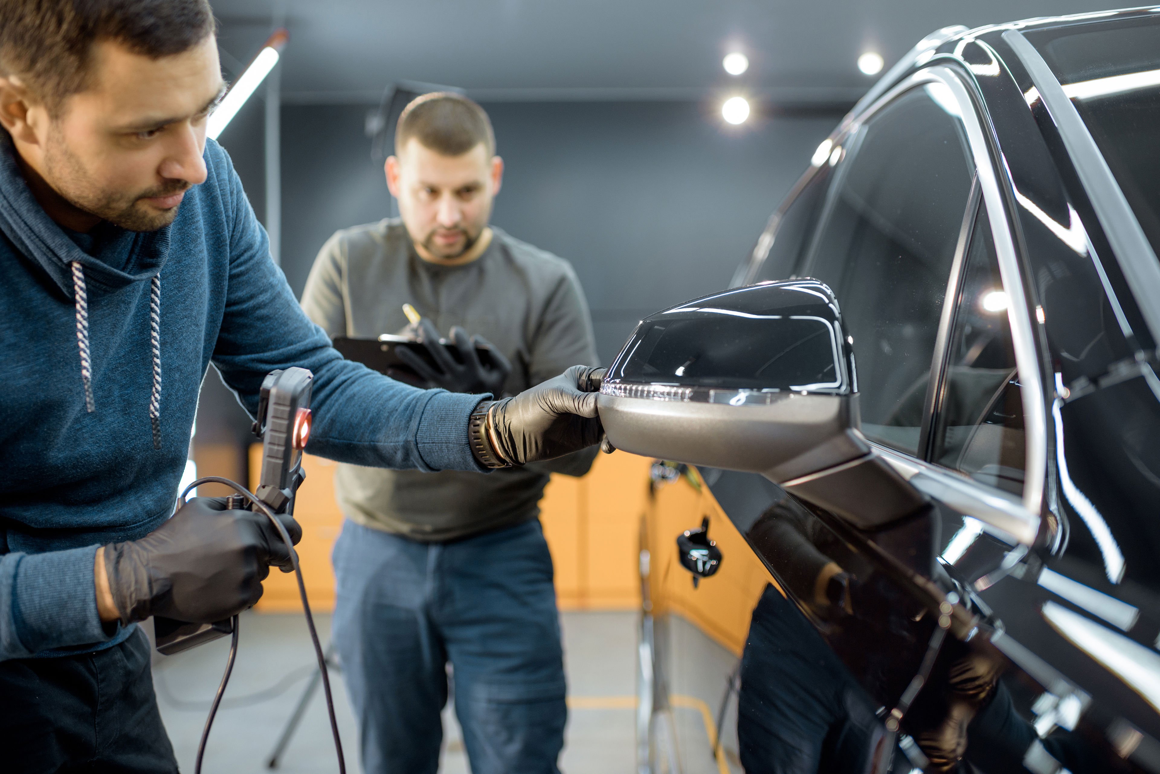 Two male technicians examining the finish on the left rear-view mirror on a black passenger car