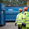 Two recycling experts dressed in hardhats and reflective Stena Recycling work clothes standing beside a couple of large blue recycling containers in a recycling yard.