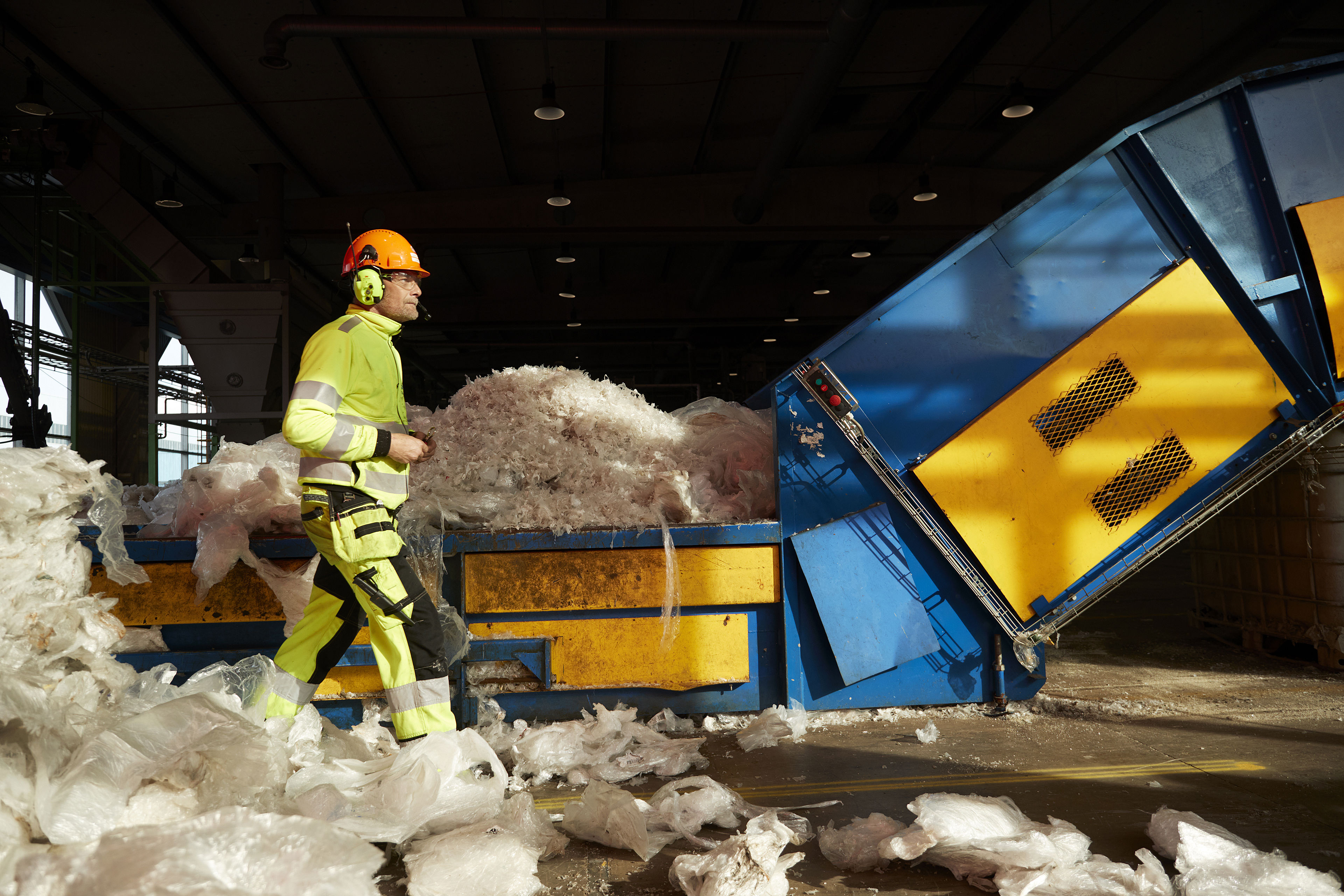 Specjalista ds. recyklingu ubrany w odblaskową odzież roboczą, w kasku, ochronnikach słuchu i okularach ochronnych, przechodzący obok maszyny do recyklingu plastiku.