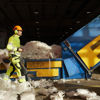 Recycling expert dressed in reflective workwear, wearing a helmet, hearing protection and safety glasses, walking past a machine that recycles plastic.