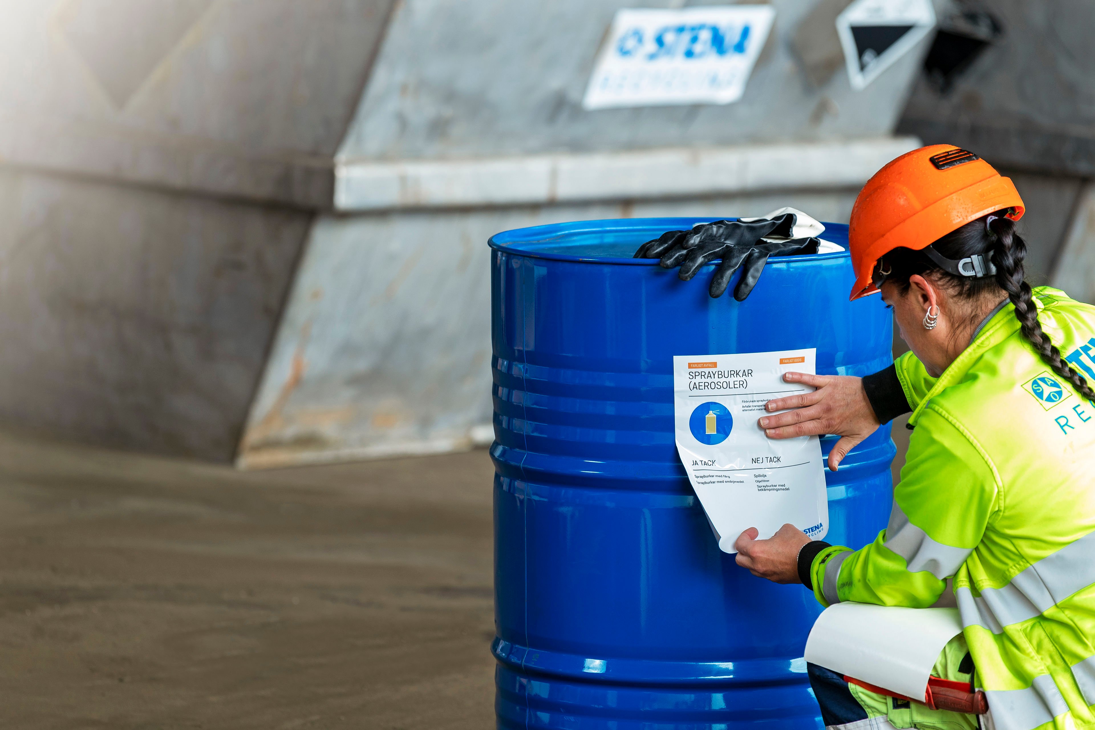 A Stena Recycling expert dressed in hardhat and reflective work jacket checking the safety label on a blue steel barrel containing hazardous waste for recycling.