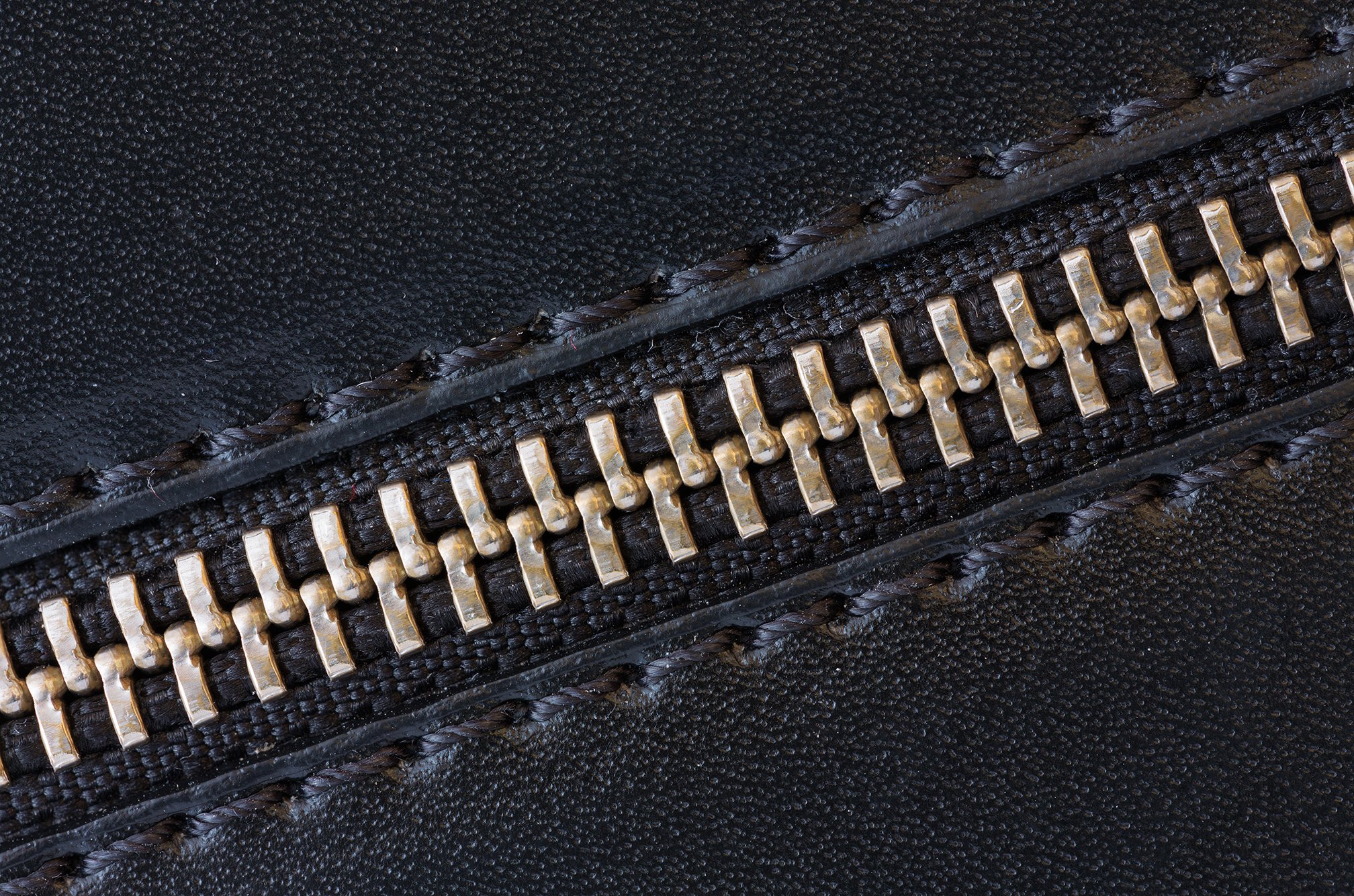A close-up image of a black faux leather texture featuring a metallic zipper with gold teeth.